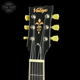 Close-up of a guitar headstock with a black finish, gold 'Vintage' script logo, a gold fleur-de-lis emblem, and light yellow tuning pegs against a black background.