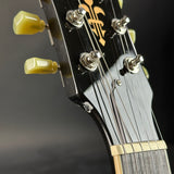 Close-up of the headstock of an acoustic guitar, showing a glossy black wood finish, gold fleur-de-lis logo, silver tuning pegs, and light yellow-green tuning keys against a dark background.