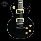 A black electric guitar with gold hardware and trim, shown from the front against a black background. A Tone Shop watermark is visible in the top-left corner.