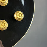 Close-up of a black electric guitar body with gold-toned control knobs and a gold edge trim, photographed against a gray background.