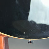 Close-up of the glossy black back of an electric guitar body, showing visible scratches and a small metal tuning peg at the bottom edge.