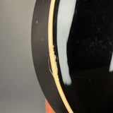Close-up of a glossy black circular object, likely a guitar body or similar instrument edge, showing a light wood rim and a small amount of loose string near the edge.