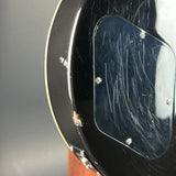 Close-up of the lower bout corner of a black electric guitar body, showing a glossy black finish, a clear pickguard with visible scratches, and a small exposed wood spot near the edge.