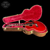 A red acoustic guitar displayed inside an open brown hard case with bright red velvet lining, photographed against a solid black background. A Tone Shop watermark is visible in the top-left corner.