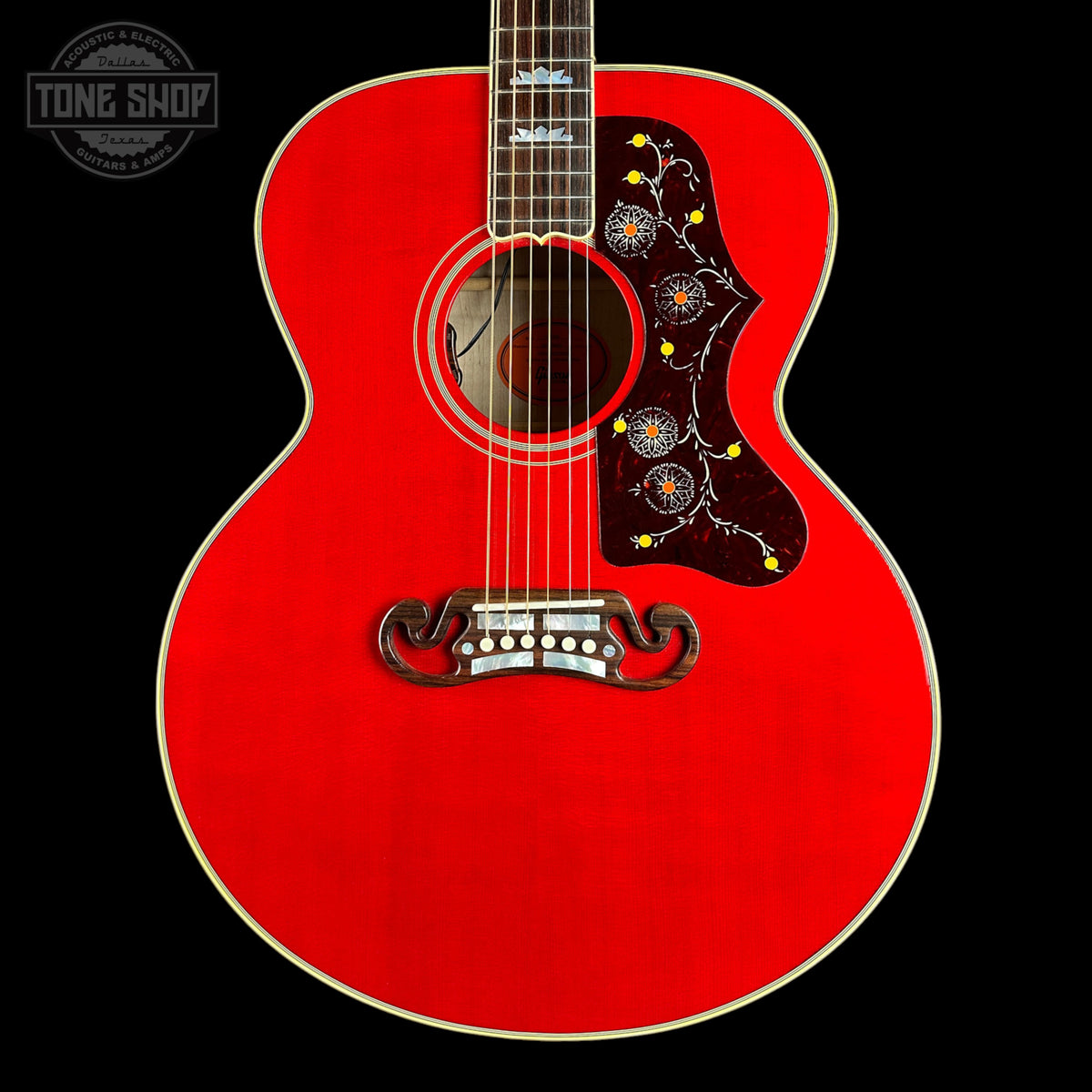 A red acoustic guitar shown from the front against a black background, featuring a decorative dark pickguard with yellow and blue floral details, a white bridge, and a white logo on the headstock partially visible at the top.