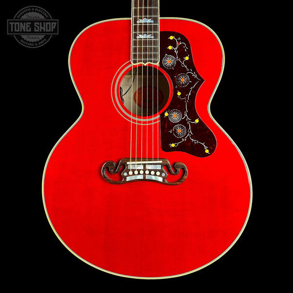 A red acoustic guitar shown from the front against a black background, featuring a decorative dark pickguard with yellow and blue floral details, a white bridge, and a white logo on the headstock partially visible at the top.