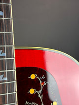 Close-up of an acoustic guitar body showing a glossy red backplate with floral inlay, a dark wood fretboard, and a dark gray background.