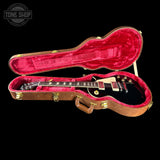 An open brown guitar hard case with a bright pink velvet interior, holding a black electric guitar. The case has gold-tone latches and a handle, and the guitar has gold hardware.