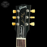 Close-up of a Gibson Les Paul Standard electric guitar headstock, featuring a black finish, gold tuning pegs, and white and gold branding text, set against a solid black background.