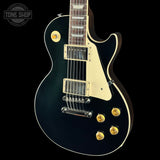 A black electric guitar with gold hardware and a cream pickguard, shown from a front three-quarter angle against a black background. A Tone Shop watermark is visible in the top-left corner.