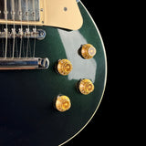 Close-up of an electric guitar body showing a dark green metallic finish, gold control knobs, a cream pickguard, and part of the bridge against a black background.