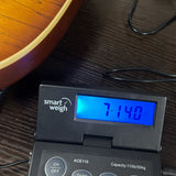A close-up of a black smart weigh electronic scale displaying 7.140 kg on its blue backlit LCD screen, resting on a dark wood tabletop next to a guitar.
