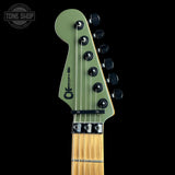 Close-up of the headstock of an electric guitar with an olive green finish, black tuning pegs, and a maple neck, shown against a solid black background. A Tone Shop watermark is visible in the top left corner.