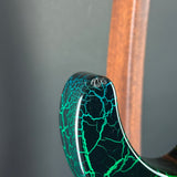 Close-up of a glossy black guitar body with bright green crackled vein pattern, showing a small clear dome pickup mount on the upper bout, with a wooden neck stock in the background.