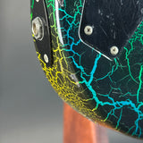 Close-up of an electric guitar body showing a glossy black finish with a vibrant green and yellow lightning-bolt crackle pattern, mounted on a wooden stand against a blurred gray background.