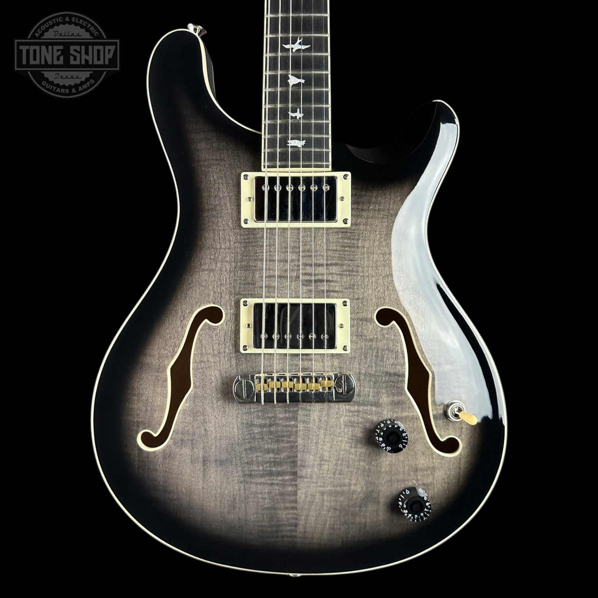 A close-up of a double-cutaway electric guitar against a solid black background. The guitar features a dark brown to black flamed maple top with a sunburst gradient, gold hardware, and white binding.