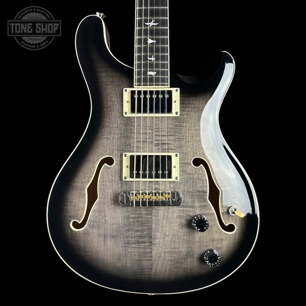 A close-up of a double-cutaway electric guitar against a solid black background. The guitar features a dark brown to black flamed maple top with a sunburst gradient, gold hardware, and white binding.
