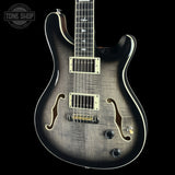 A dark gray to black flamed maple electric guitar with gold hardware and white binding, shown against a solid black background with a Tone Shop watermark in the top left corner.