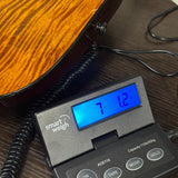 A close-up of a digital smart scale labeled 'smart weigh ACE110' on a dark wooden surface, next to the body of an electric guitar. The scale's blue backlit display is active, showing weight readings.