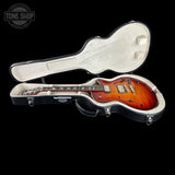 A sunburst electric guitar shown inside an open black hard case with cream lining, photographed on a black background. A Tone Shop watermark appears in the upper left corner.