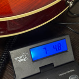 A close-up of a digital smart weigh scale displaying '7.48 kg' on a dark wooden surface, with part of a red electric guitar visible at the top.