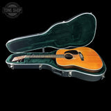 An acoustic guitar with a natural wood finish resting inside an open black hard case with a green velvet interior, shown against a solid black background. A Tone Shop watermark is visible in the top-left corner.