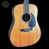 An acoustic guitar with a natural wood finish and a black pickguard, shown against a solid black background with a Tone Shop watermark in the top left corner.