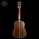 The back of a wooden acoustic guitar, showing a rich dark brown wood grain finish, a central soundhole, and a six-string headstock, photographed against a solid black background with a Tone Shop watermark in the top left corner.