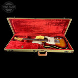 A classic electric guitar with a sunburst finish and white pickguard resting inside an open vintage hard case with a red velvet interior, set against a solid black background.
