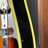 Close-up of a guitar body showing a black glossy finish with a yellow binding edge, part of a metal headstock, and a wood grain surface in the background.