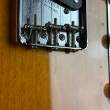 Close-up of a guitar's tremolo bridge and tailpiece hardware, mounted on a sunburst wooden body. The metal bridge assembly has visible wear and scratches, and the wood grain is prominent.