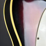 Close-up of a glossy sunburst electric guitar body showing the dark center, amber-red outer edge, gold binding, and part of a white pickguard.