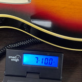 A close-up of a guitar body with a sunburst finish and cream binding, resting on a dark wooden surface next to a digital smart weigh scale displaying 7.100 oz.