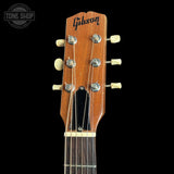 Close-up of a Gibson acoustic guitar headstock with six cream-colored tuning pegs and strings, set against a solid black background.