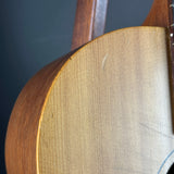 Close-up detail of an acoustic guitar's wooden body, showing the natural wood grain, soundboard, and part of the neck against a dark background.