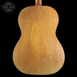 The back of a vintage acoustic guitar made of natural wood, showing a warm brown grain and wear marks, set against a solid black background with a Tone Shop watermark in the top left.