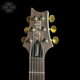 Close-up of the headstock of a Takamine acoustic guitar against a black background, featuring a dark wood grain finish, gold tuning pegs, and a white signature on the backplate.