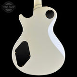 The back of an electric guitar body in a glossy off-white finish, featuring black pickguard areas with gold screws, shown against a solid black background.