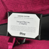 A close-up of a Gibson Custom Certificate of Authenticity card placed on a black case, set against rich magenta-pink fabric with a matching ribbon.