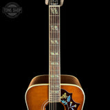 Close-up of the neck and lower body of an acoustic guitar against a black background, showing a natural wood finish, decorative inlay on the fretboard, and floral inlay on the lower bout.