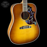 A close-up of an acoustic guitar body with a natural sunburst finish, featuring intricate blue and white floral inlay on the pickguard and a white binding around the body edge, set against a solid black background.