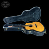An acoustic guitar with a natural wood finish resting inside an open black hard case on a solid black background. A circular 'TONE SHOP' watermark is visible in the top-left corner.