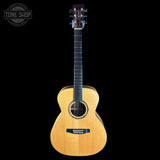 A natural wood acoustic guitar with a dark headstock and black bridge, shown vertically against a solid black background. A circular 'TONE SHOP' watermark is visible in the top-left corner.