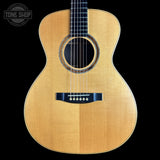 A front-facing acoustic guitar with a natural wood finish and black hardware, shown against a solid black background with a Tone Shop logo in the top-left corner.
