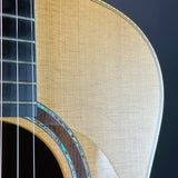 Close-up detail of an acoustic guitar body showing the natural wood grain, sound hole rosette with iridescent inlay, and part of the dark fretboard and strings against a dark background.