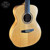 An acoustic guitar with a natural wood finish and black hardware, shown against a solid black background with a Tone Shop logo in the top-left corner.
