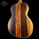 Close-up of the back of an acoustic guitar showing a rich wood grain pattern with vertical stripes of dark brown, medium brown, and warm honey tones, finished with a light natural wood edge and neck against a solid black background.