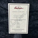 A vintage-style Jackson Custom Shop factory certificate card with black text on an off-white textured background, placed on a dark plush fabric surface.