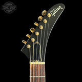 Close-up of the headstock of a Gibson electric guitar against a solid black background, showing the Gibson logo, gold tuning pegs, and a dark wood fretboard.