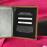 An open Gibson Custom Certificate of Authenticity booklet on a bright pink fabric background. The black insert shows white text and handwritten name and number labels.